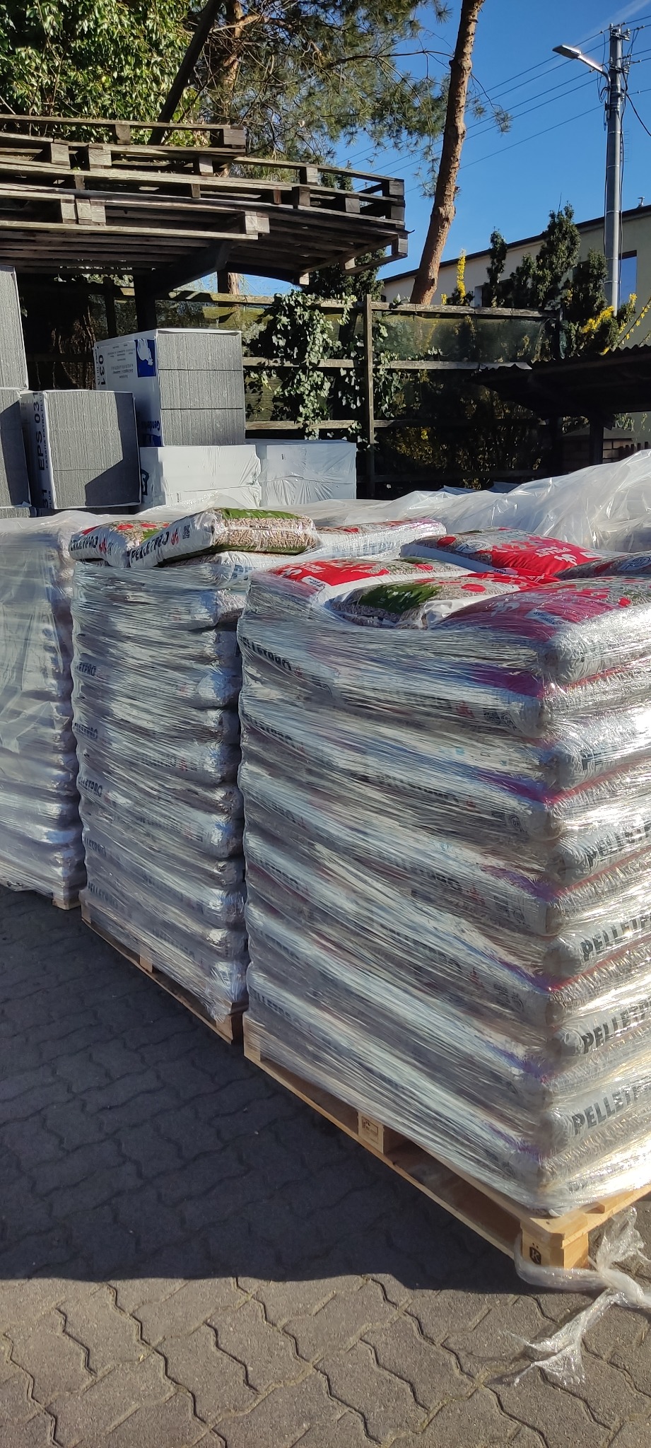 Składowane palety z workami pelletu, owinięte folią, na zewnątrz. Widoczne drzewa i budynek w tle. Ujęcie w słoneczny dzień.