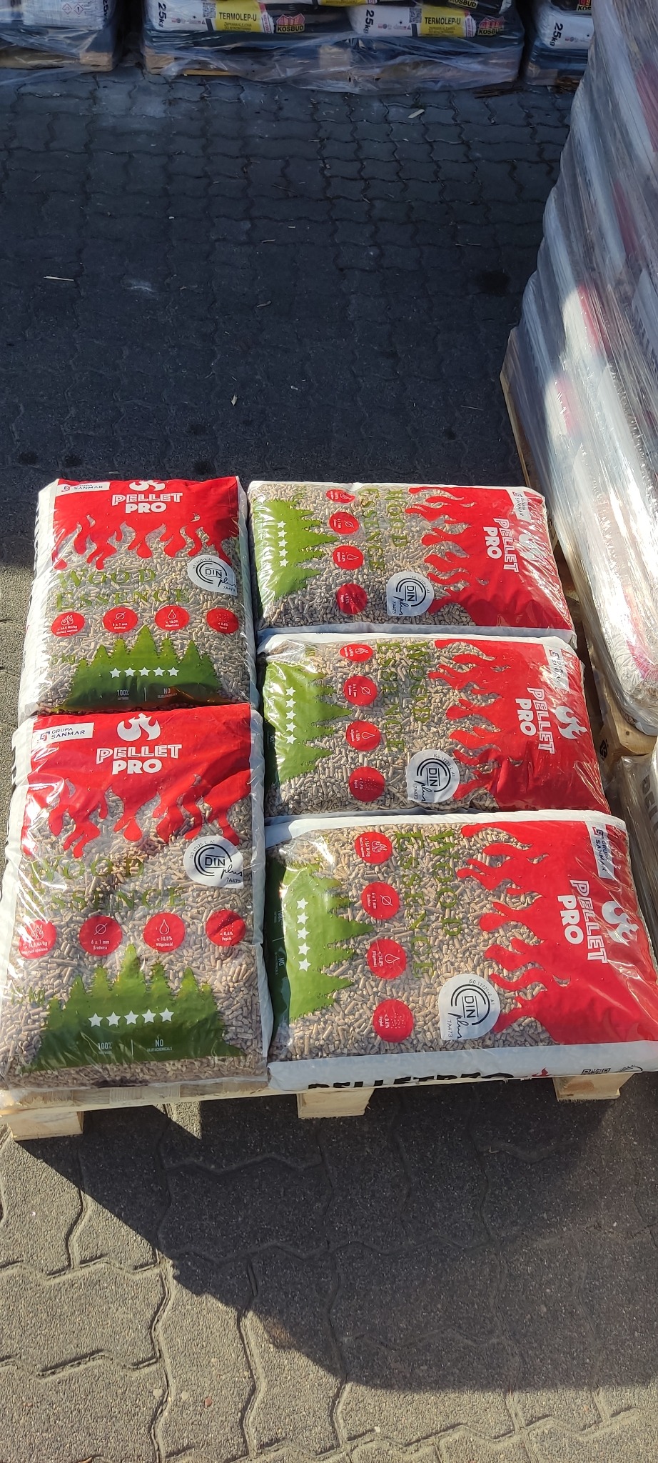 Ułożone na palecie worki z pelletem drzewnym, marki Pellet Pro, z widocznymi certyfikatami DIN Plus, w otoczeniu innych zapakowanych materiałów budowlanych.