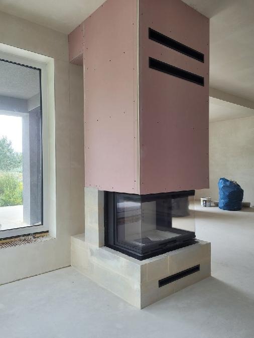 Wnętrze z kominkiem w trakcie zabudowy, obudowa z płyt karton-gips, widoczne otwory wentylacyjne, surowy stan wykończenia, okno z widokiem na zieleń.