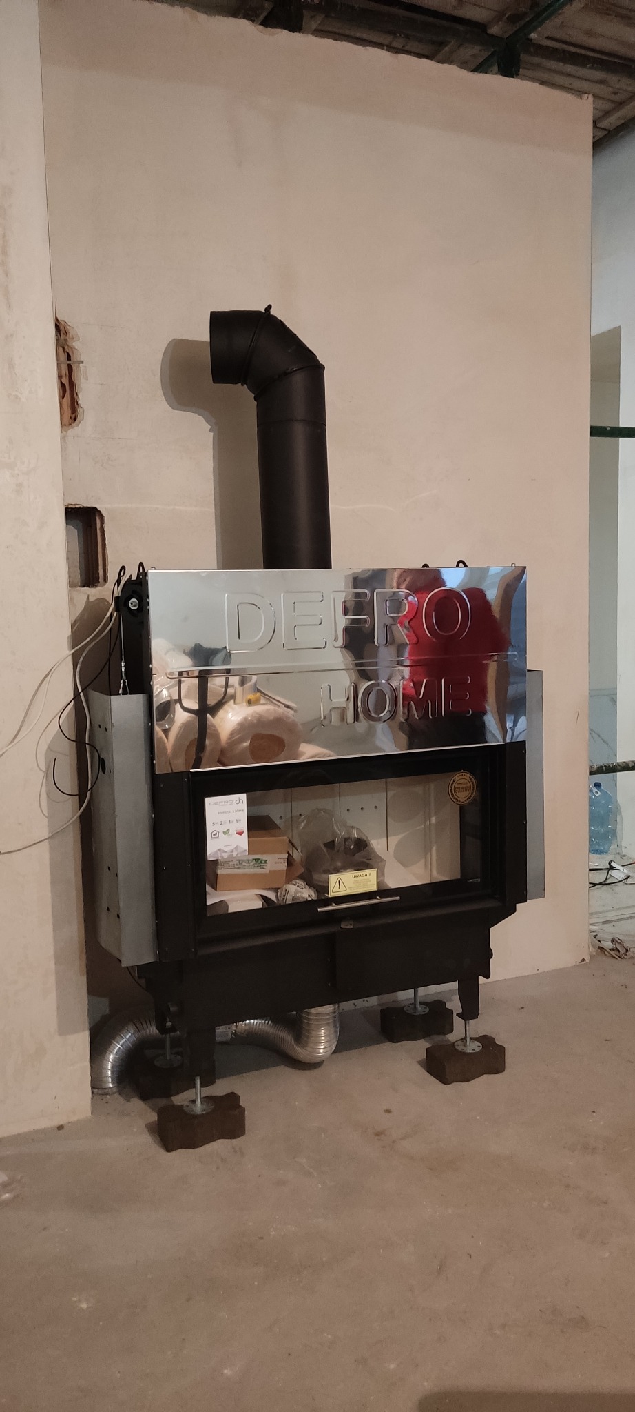 Nowoczesny piec na pellet marki Defro Home, z widocznym kominem i elementami instalacji, ustawiony w surowym wnętrzu z nieotynkowanymi ścianami.