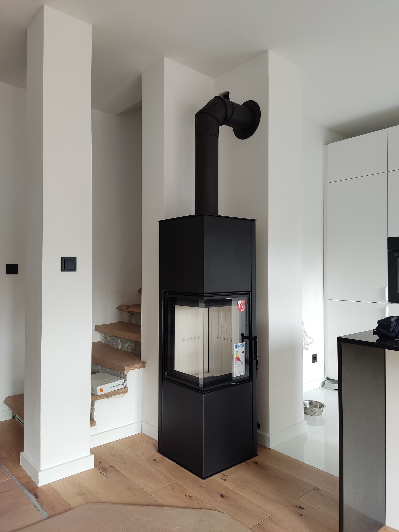 Nowoczesny, czarny piec wolnostojący z szybą, z podłączonym kominem. Wnętrze w bieli, schody z drewnianymi stopniami. Podłoga z jasnych desek.