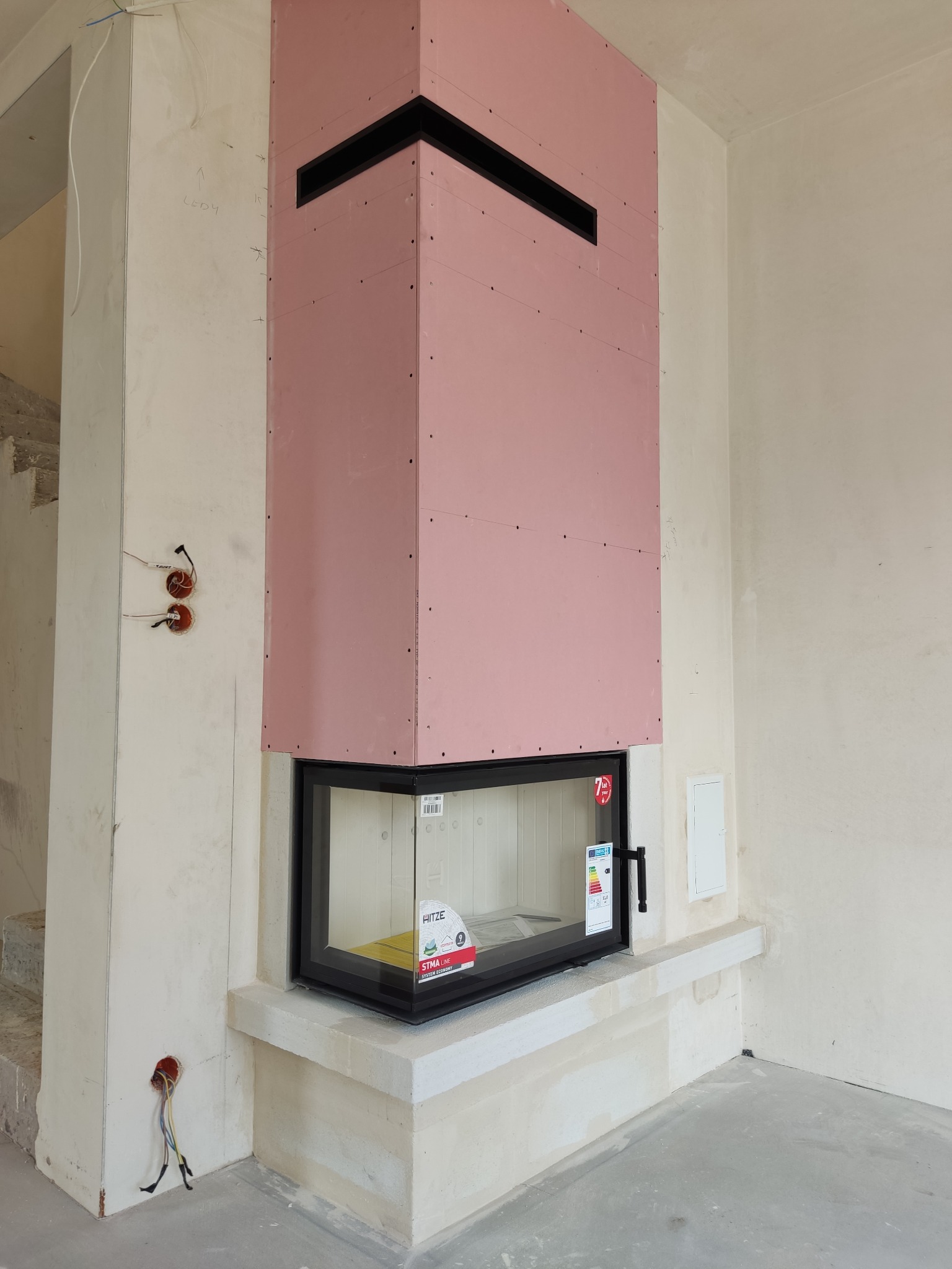 Wnętrze w trakcie budowy: obudowa kominka z różowych płyt kartonowo-gipsowych z czarnymi wlotami powietrza, osadzona na surowym, betonowym cokole.