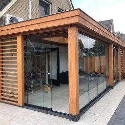 Aluwooden - Zabudowa tarasu z drewna i szkła, z przesuwnymi panelami szklanymi i drewnianymi lamelami, widok z zewnątrz na jasną podłogę i część domu.