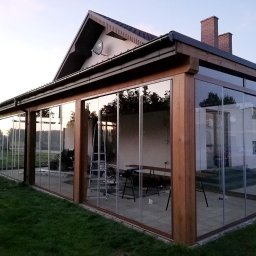 Aluwooden - Szklana zabudowa tarasu z drewnianą konstrukcją. Widoczne przesuwne szklane panele i fragment domu z ceglanym kominem. Na tarasie drabina i stół.
