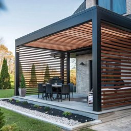 Aluwooden - Nowoczesna pergola z czarną ramą i drewnianymi lamelami, z zestawem mebli ogrodowych, na tle zadbanego ogrodu i domu z cegły.