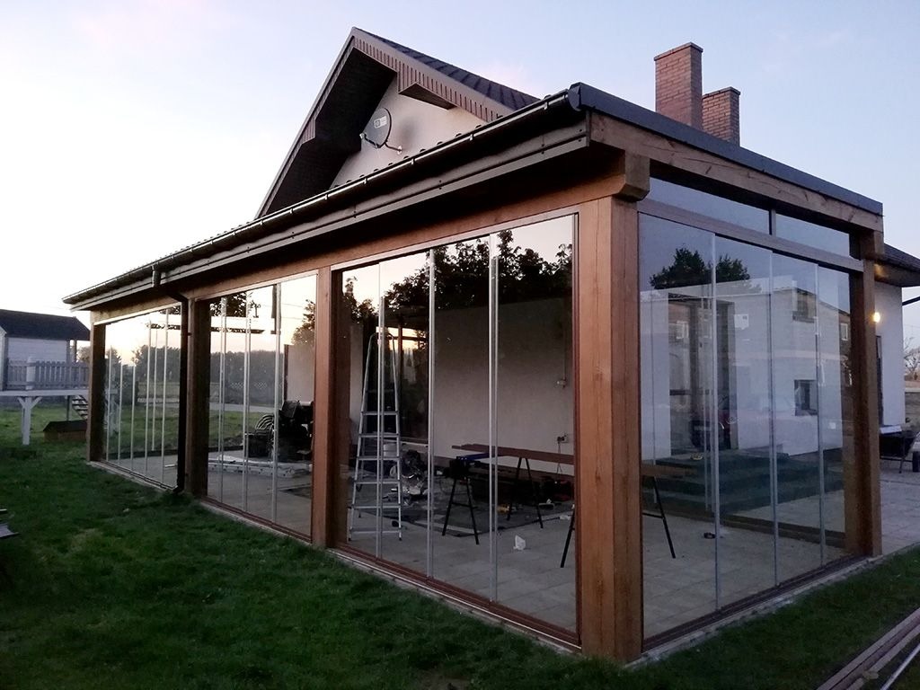 Szklana zabudowa tarasu z drewnianą konstrukcją. Widoczne przesuwne szklane panele i fragment domu z ceglanym kominem. Na tarasie drabina i stół.