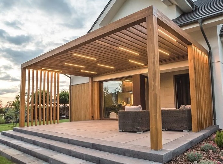 Drewniana pergola z oświetleniem LED nad tarasem z szarych płyt. Pionowe drewniane panele zapewniają częściową osłonę. Nowoczesny design z elementami naturalnego drewna.