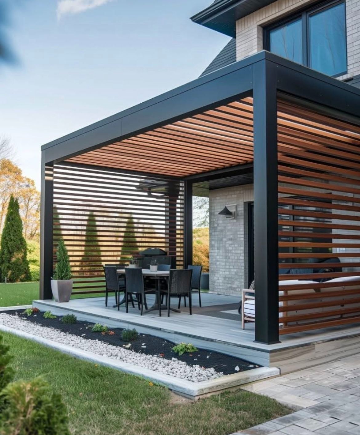 Nowoczesna pergola z czarną ramą i drewnianymi lamelami, z zestawem mebli ogrodowych, na tle zadbanego ogrodu i domu z cegły.