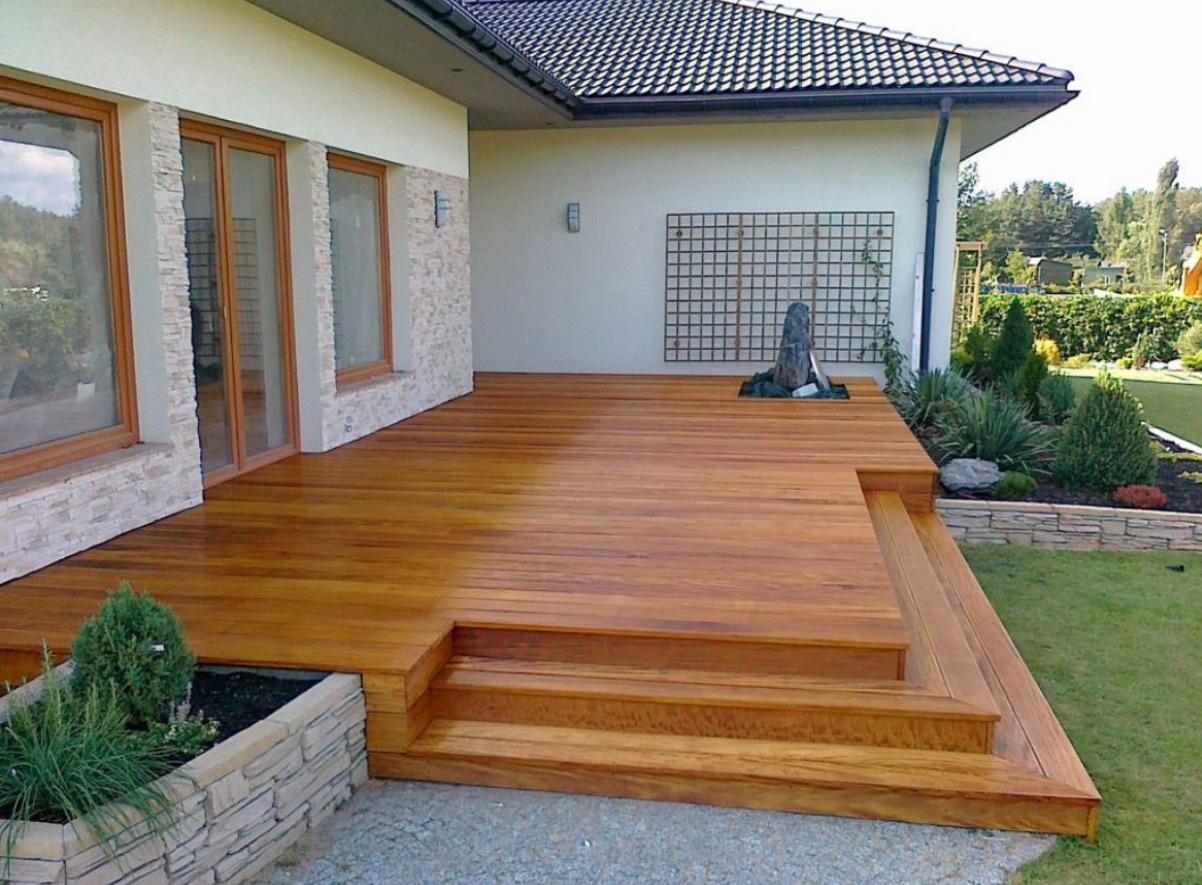 Elegancki taras drewniany z egzotycznego drewna przy domu. Widoczne schody, donica z kamienia i ozdobna krata z elementami roślinnymi na ścianie.