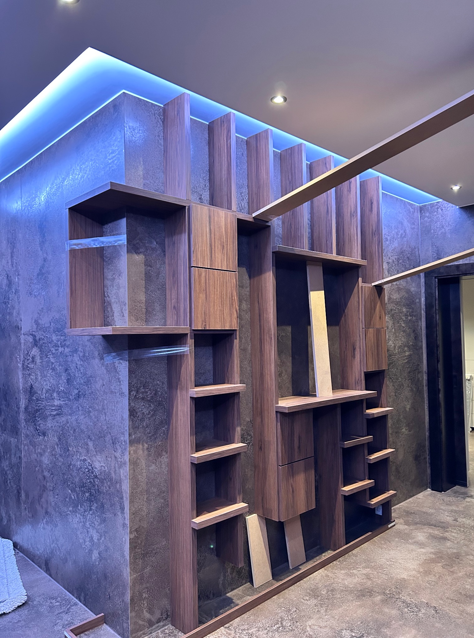 Nowoczesna szafa wnękowa z ciemnego drewna, podświetlona niebieskim światłem LED, z widocznymi elementami konstrukcyjnymi i surowym wykończeniem ściany.