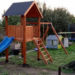 Drewniany plac zabaw z niebieską zjeżdżalnią i huśtawką na tle zielonego ogrodu z trampoliną i domkiem dla dzieci. Widoczne drzewa i krzewy.