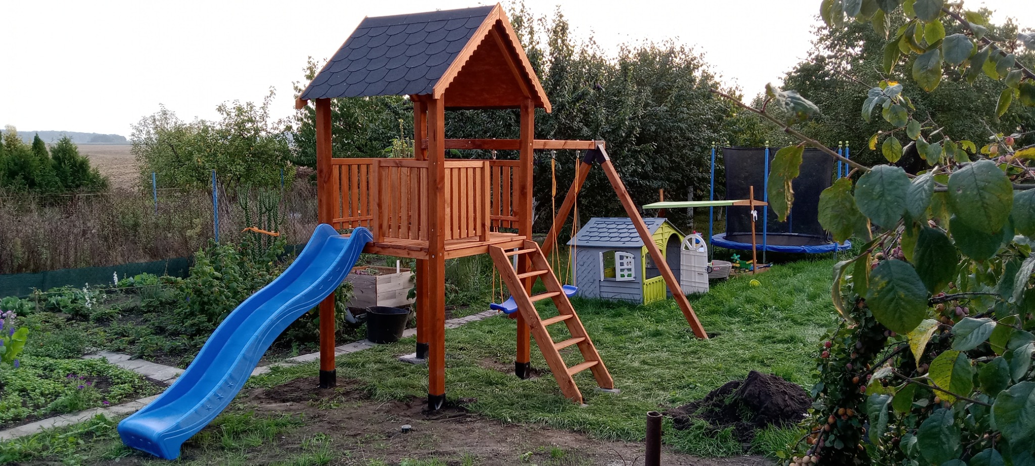 Drewniany plac zabaw z niebieską zjeżdżalnią i huśtawką na tle zielonego ogrodu z trampoliną i domkiem dla dzieci. Widoczne drzewa i krzewy.