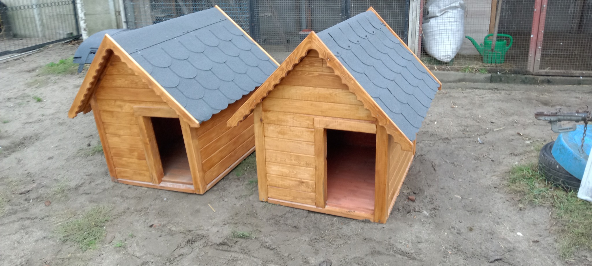 Dwie drewniane budy dla psów z szarymi dachami, ustawione na ziemi. Widoczne detale wykończenia i faktura drewna. W tle siatka ogrodzeniowa i akcesoria ogrodowe.