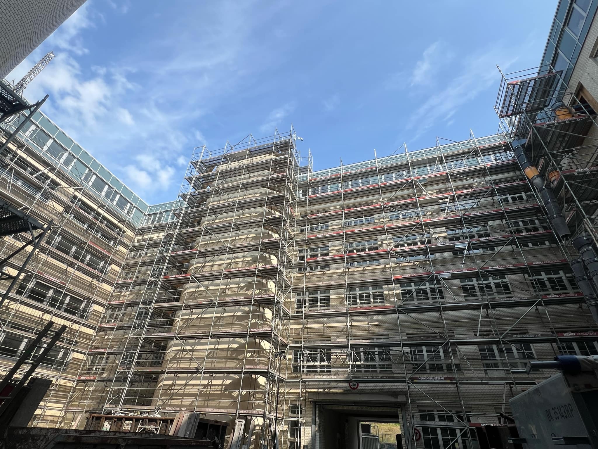 Budynek w rusztowaniach, widok od dołu, przygotowania do prac elewacyjnych. Na rusztowaniach widoczne tabliczki z nazwą firmy. Błękitne niebo w tle.