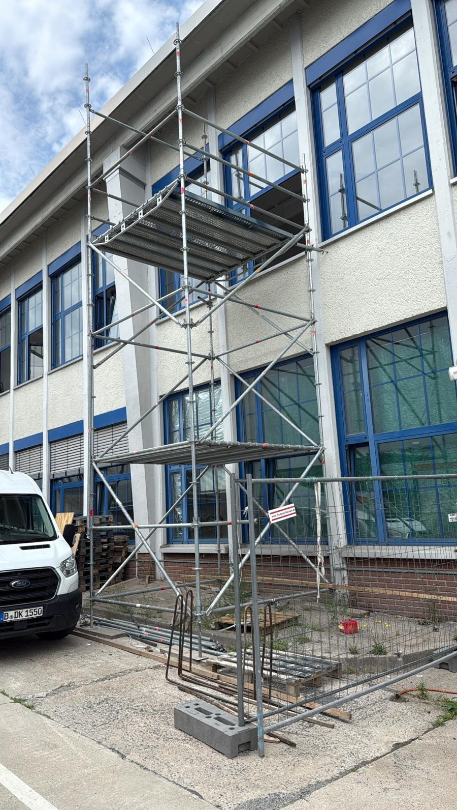 Metalowe rusztowanie przy budynku z niebieskimi oknami, widoczny fragment białego busa. Prace na wysokościach, zabezpieczenie terenu siatką.
