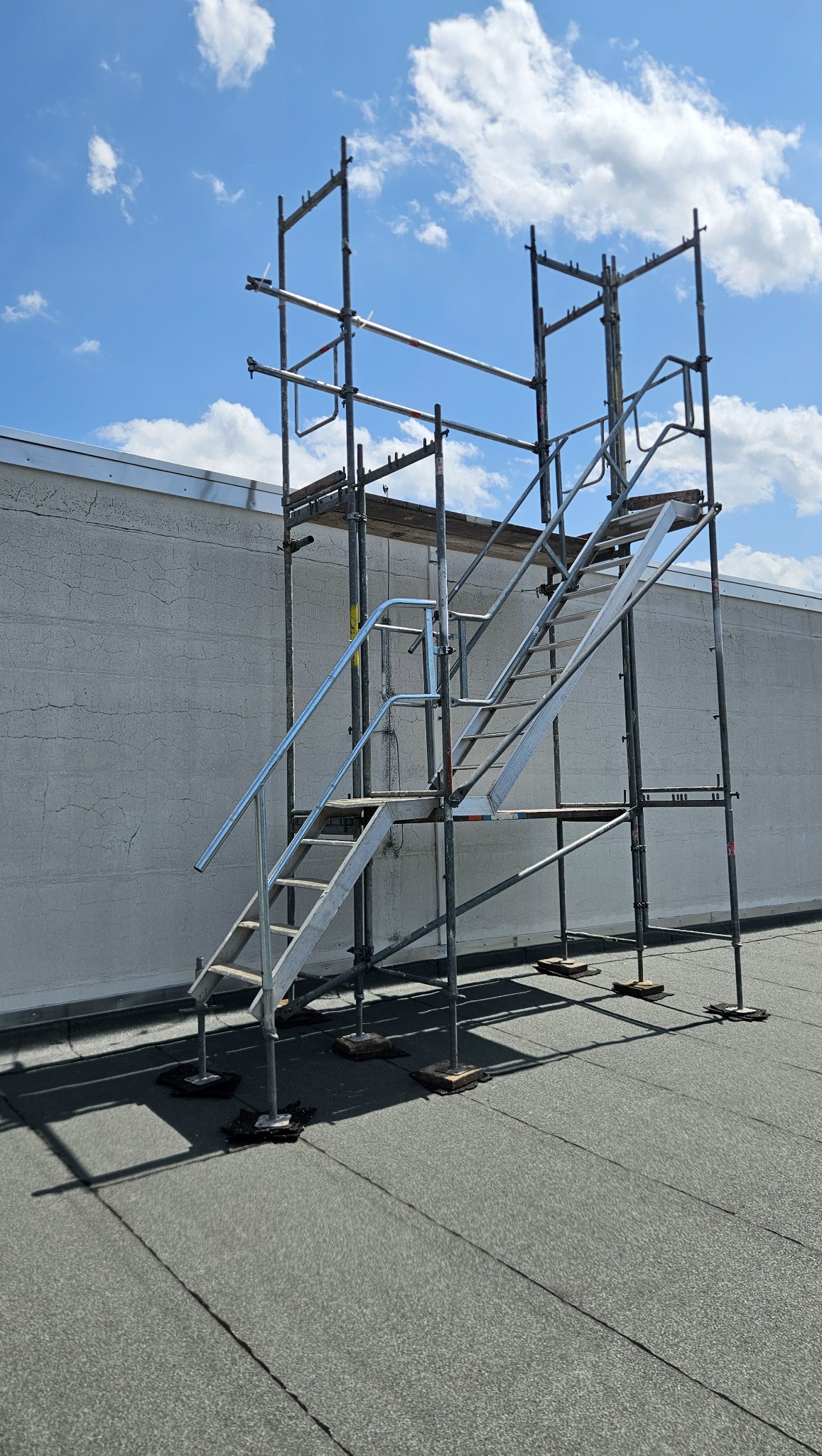 Metalowe rusztowanie schodowe na dachu budynku, widoczne na tle nieba z chmurami. Konstrukcja wsparta na regulowanych podstawach, rzucająca cień na pokrycie dachowe.