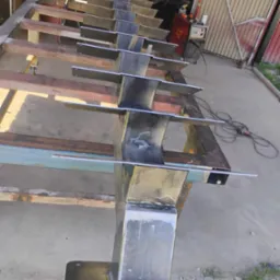 Metalowa konstrukcja w trakcie spawania, widoczne spawy i elementy mocujące. Konstrukcja umieszczona na metalowym stole warsztatowym, w tle sprzęt spawalniczy.