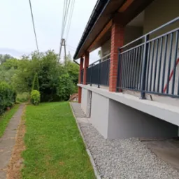 Nowoczesna balustrada w kolorze antracytu, zamontowana na balkonie domu z ceglanymi filarami, widok z boku na tle zielonego ogrodu i ścieżki.