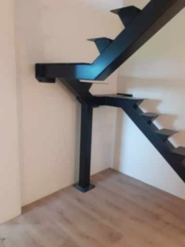 Nowoczesne, czarne schody metalowe wspornikowe, przymocowane do ściany, z widoczną konstrukcją nośną i drewnianą podłogą. Minimalistyczny design.