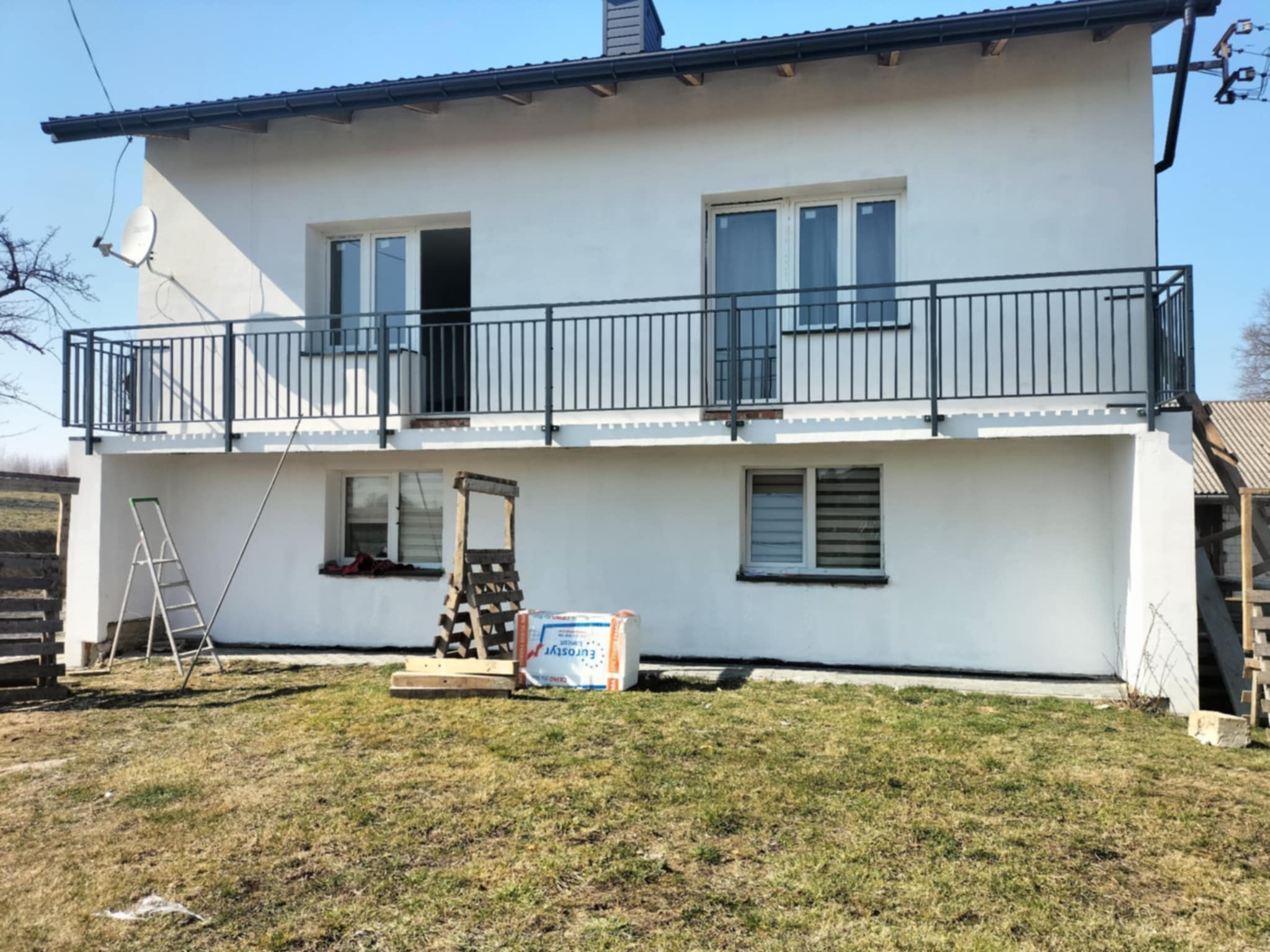 Biały dom z ciemną balustradą balkonową i drabina oparta o ścianę. Na trawniku materiały budowlane, słoneczny dzień.