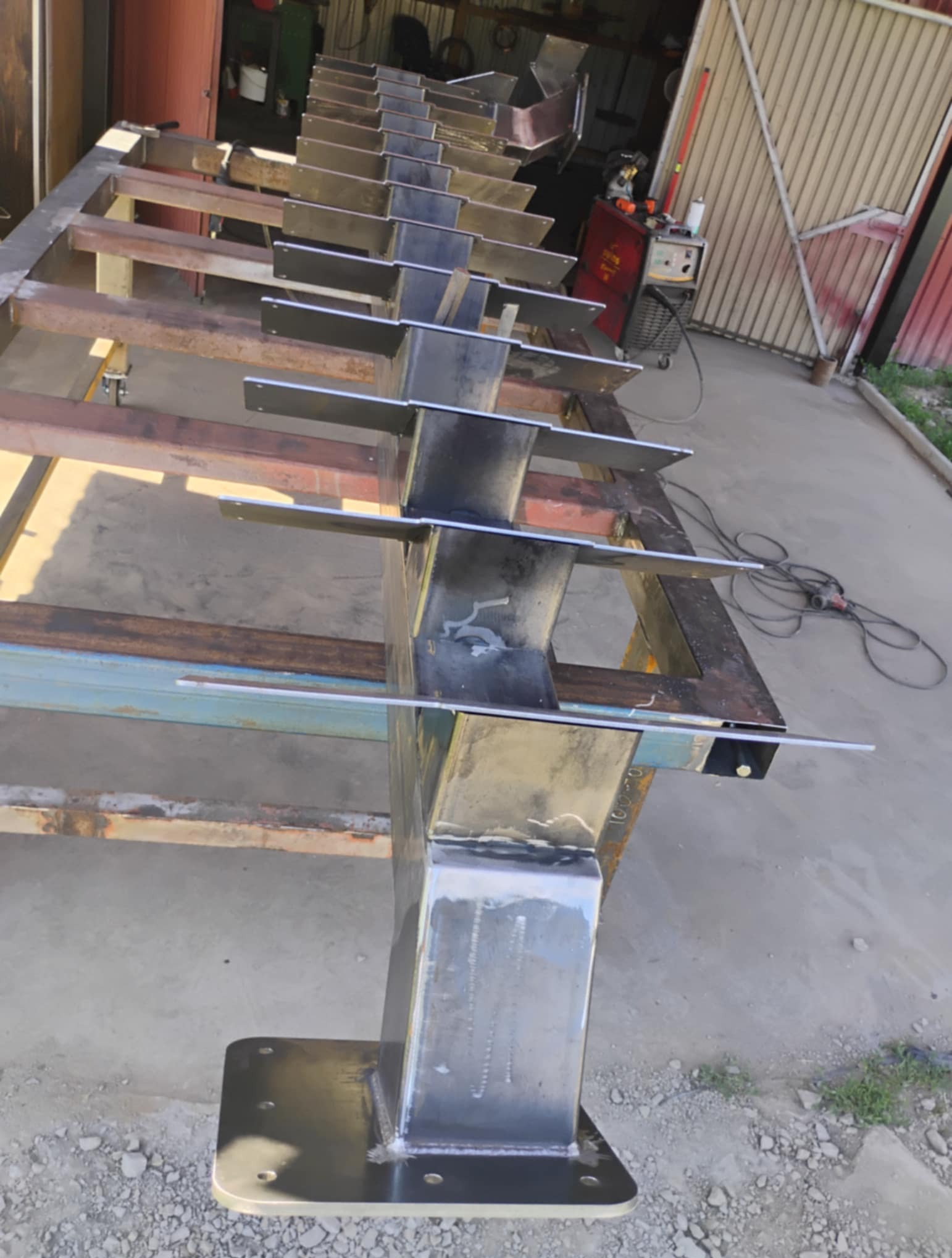 Metalowa konstrukcja w trakcie spawania, widoczne spawy i elementy mocujące. Konstrukcja umieszczona na metalowym stole warsztatowym, w tle sprzęt spawalniczy.