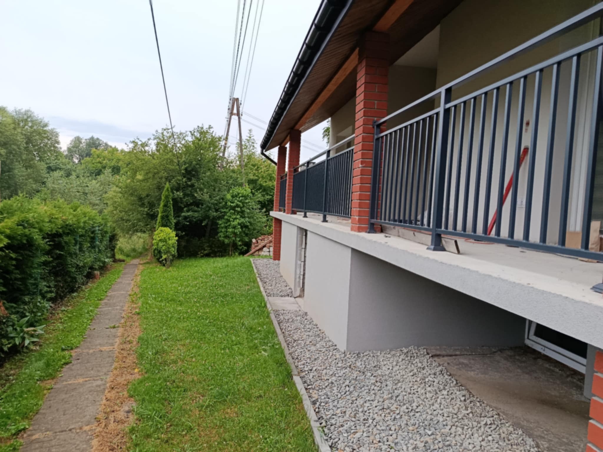 Nowoczesna balustrada w kolorze antracytu, zamontowana na balkonie domu z ceglanymi filarami, widok z boku na tle zielonego ogrodu i ścieżki.