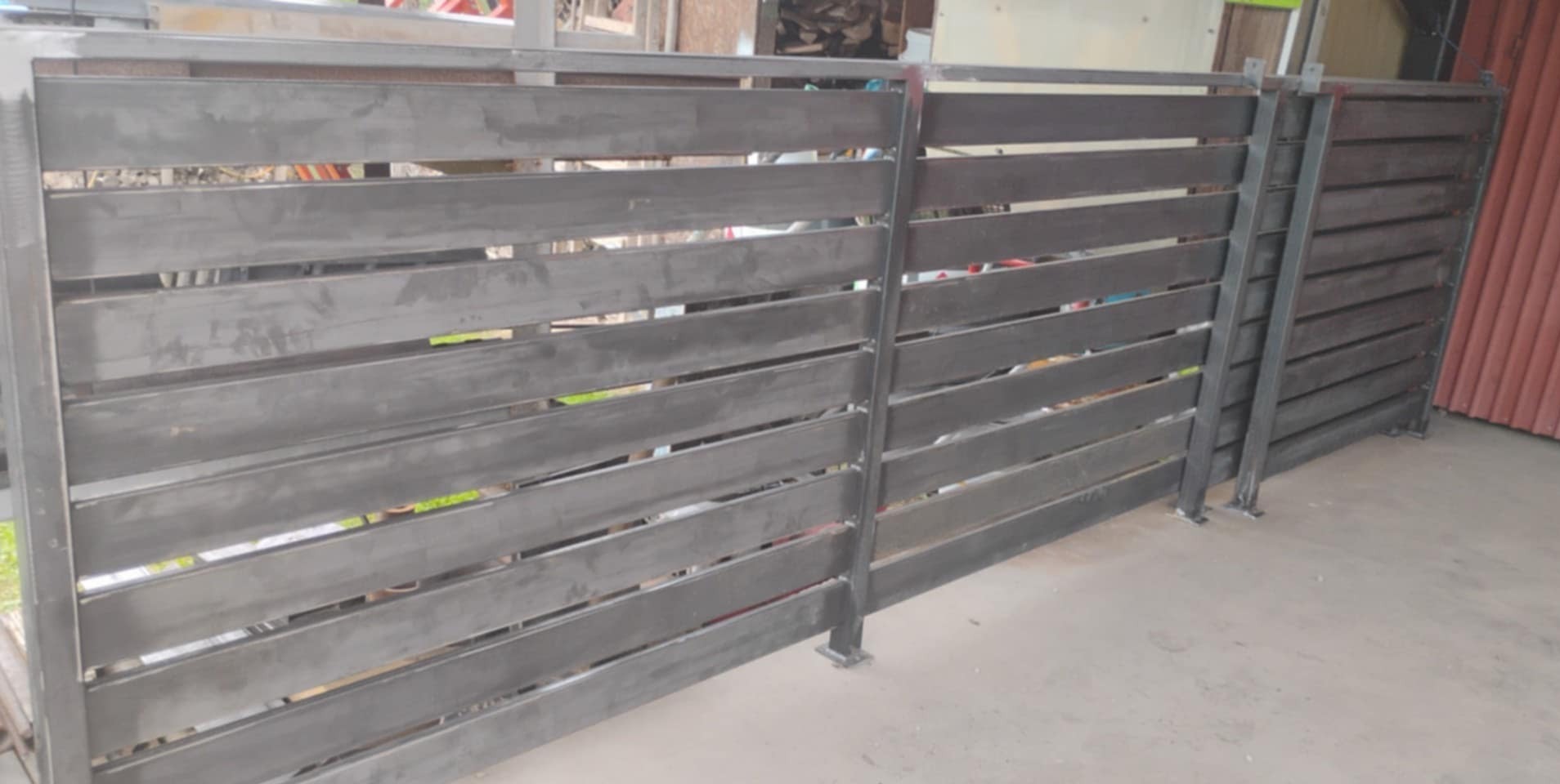 Nowoczesne, poziome ogrodzenie metalowe w surowym wykończeniu, z widocznymi spawami i minimalistycznym designem. Elementy mocujące widoczne u dołu.