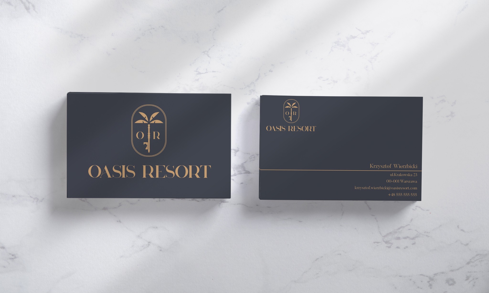 Dwie wizytówki firmy Oasis Resort z logo palmy i złotym tekstem na ciemnym tle, leżące na marmurowej powierzchni w miękkim świetle.