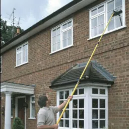 Mężczyzna czyści okna dwupiętrowego domu z cegły za pomocą długiego, żółtego wysięgnika z zamontowaną szczotką, widoczne kolumny wejściowe i antena na dachu.