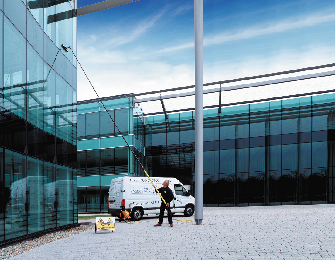 Mężczyzna myje szklaną fasadę biurowca za pomocą lancy teleskopowej z dyszą, obok zaparkowany biały bus z logo firmy czyszczącej, tabliczka ostrzegawcza o mokrej nawierzchni.