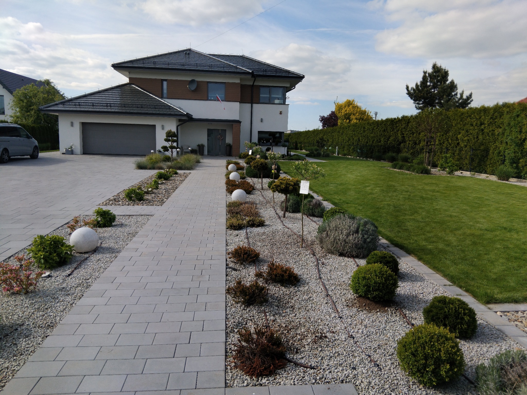 Nowoczesny dom z zadbanym ogrodem: ścieżka z kostki brukowej, ozdobne krzewy, trawnik i wysoki żywopłot. Widok na budynek z garażem i elementami dekoracyjnymi.