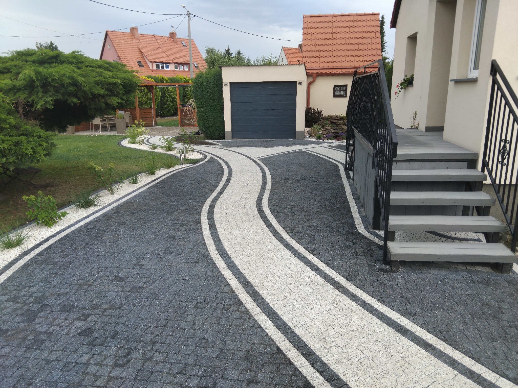 Wzorzysty podjazd z kostki brukowej w odcieniach szarości i bieli, prowadzący do domu i garażu. Widoczne schody wejściowe i elementy ogrodu.