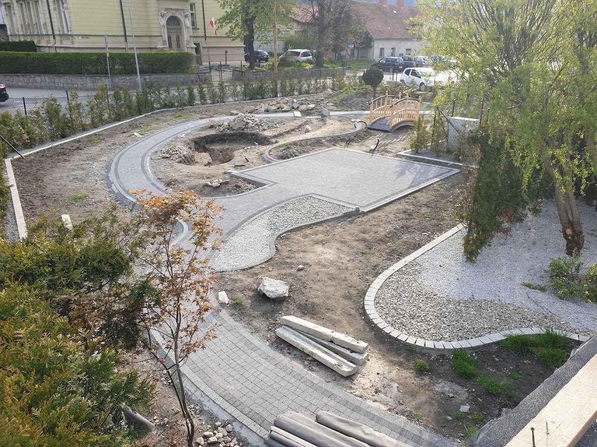 Aranżacja ogrodu w trakcie realizacji: ścieżki z kostki, obszar z kamykami, mały mostek. Widok z góry ukazuje postęp prac i elementy dekoracyjne.