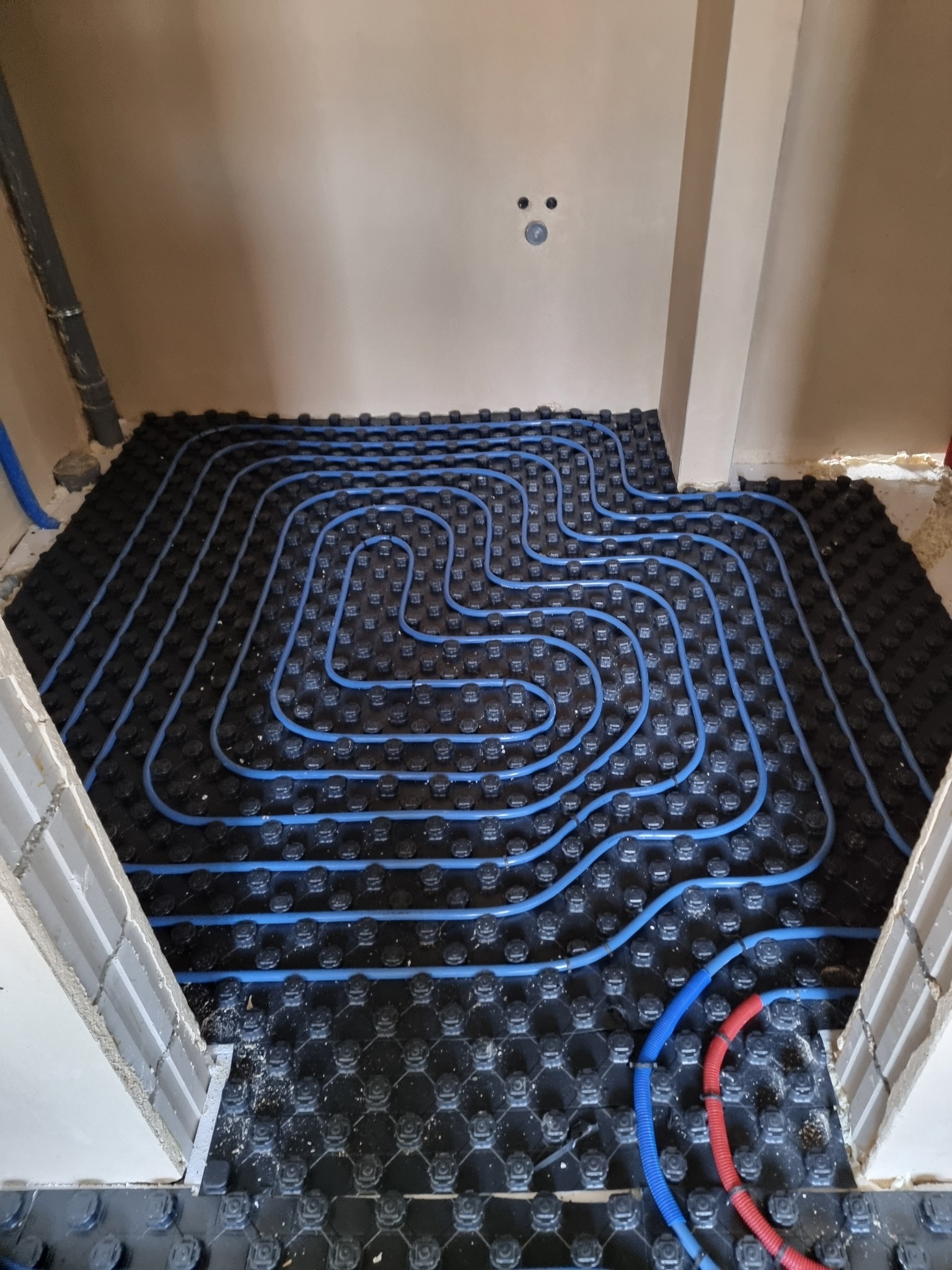 Niskie ujęcie na rozłożone, niebieskie rury ogrzewania podłogowego na czarnej macie w jasnym pomieszczeniu w trakcie realizacji.
