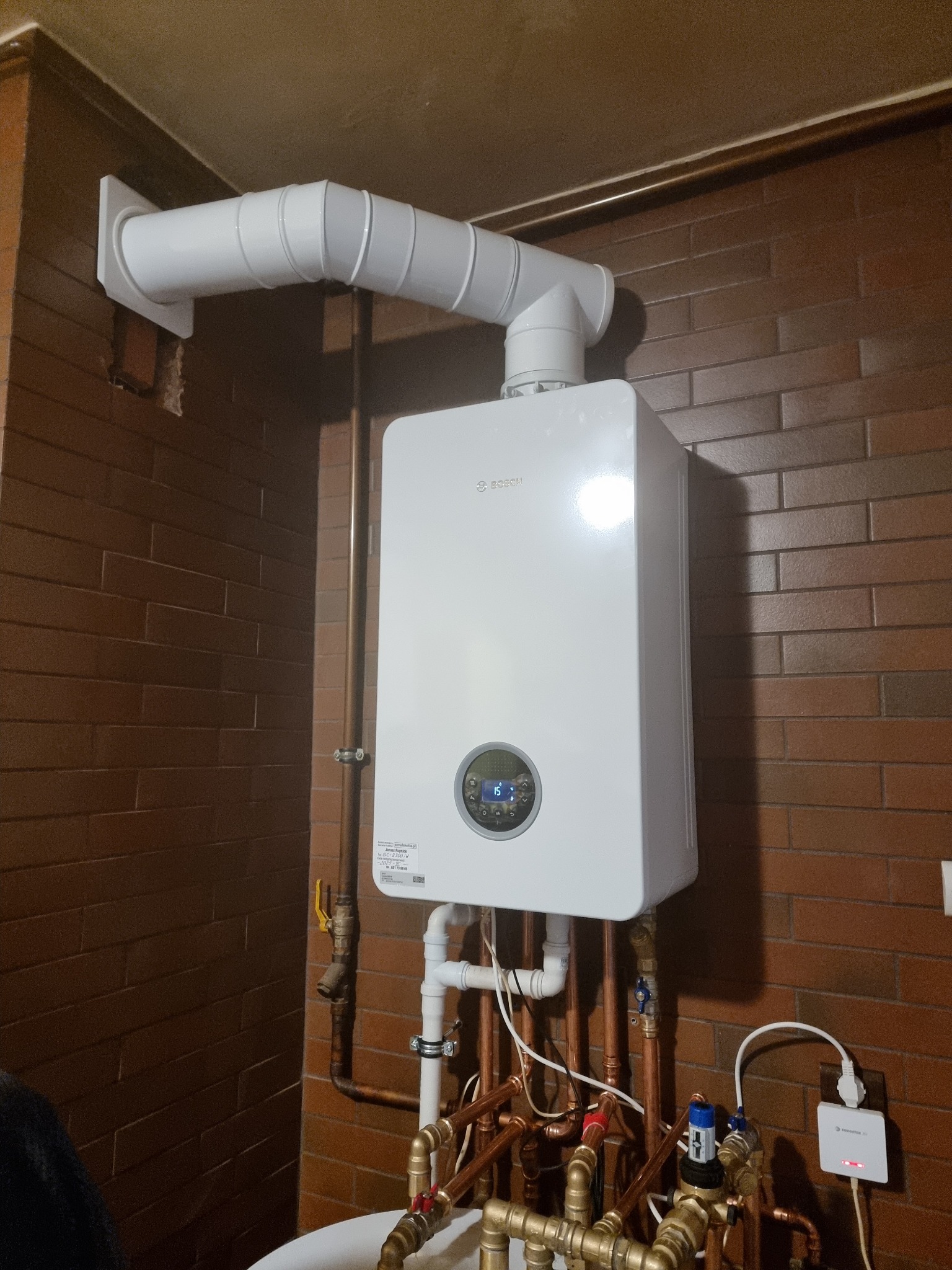 Biały piec gazowy Bosch z widocznymi miedzianymi rurami i białym kominem na tle ściany z brązowej cegły. Widoczny fragment instalacji i podłączenie do sieci.