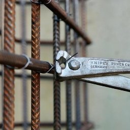 CraftBau - Zbrojenie fundamentów z użyciem metalowych prętów i narzędzia Knipex Power Cut, zbliżenie na proces wiązania drutem, szare tło.