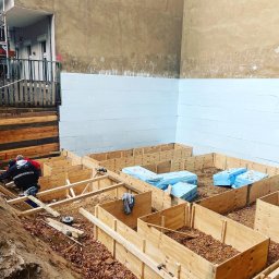 CraftBau - Budowa fundamentów: drewniane szalunki, ocieplenie styropianem, robotnicy w trakcie pracy. Widoczne narzędzia i materiały budowlane.