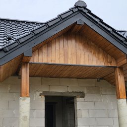 CraftBau - Zadaszenie wejścia do domu w stanie surowym: czarna dachówka, drewniana boazeria i kolumny na ścianie z białego betonu komórkowego.