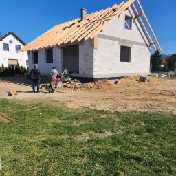 CraftBau - Budowa domu z białych bloczków: ściany wzniesione, więźba dachowa w trakcie montażu. Dwóch pracowników przy pile, przygotowuje elementy konstrukcji dachu.