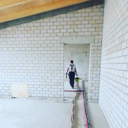 CraftBau - Wnętrze budynku w trakcie budowy: murowane ściany z białej cegły, drewniany strop i widoczne instalacje (kable, rury) na podłodze. W tle sylwetka osoby.