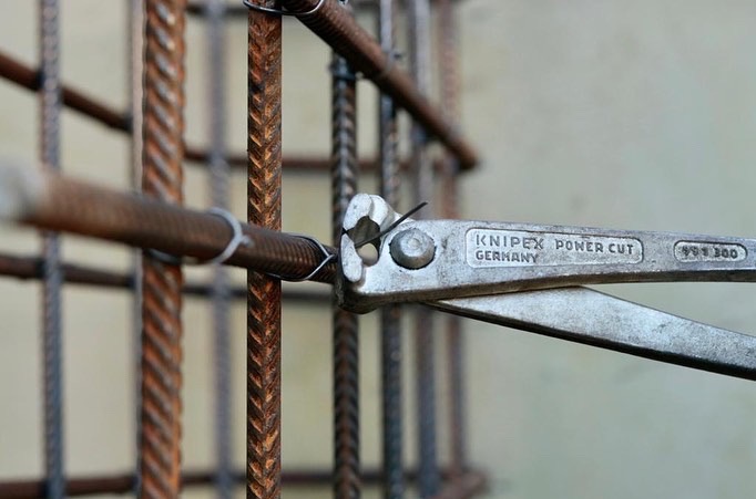 Zbrojenie fundamentów z użyciem metalowych prętów i narzędzia Knipex Power Cut, zbliżenie na proces wiązania drutem, szare tło.