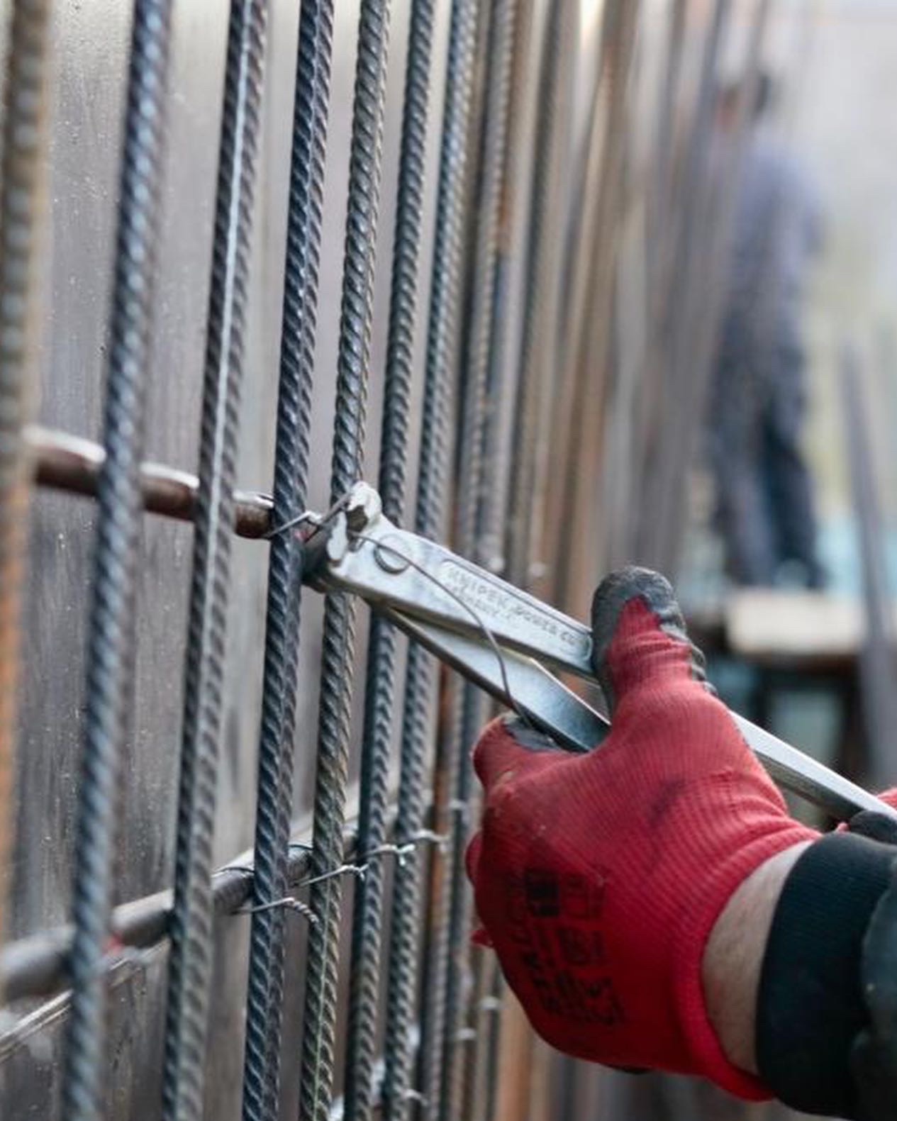 Zbrojenie pionowych prętów zbrojeniowych drutem za pomocą obcęgi Knipex, widoczne rękawice robocze i fragment sylwetki osoby w tle.