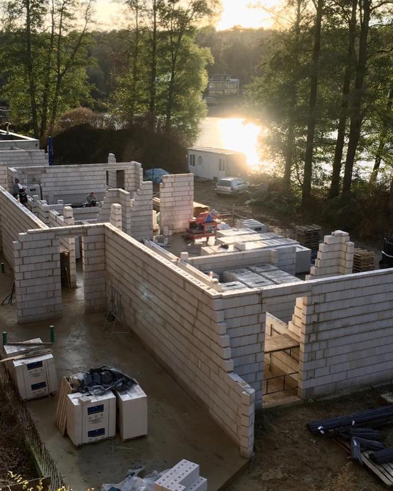 Wznoszenie ścian domu z betonu komórkowego w Szczecinie, widok z góry na budowę w otoczeniu drzew i jeziora, w tle barakowóz i samochód osobowy.