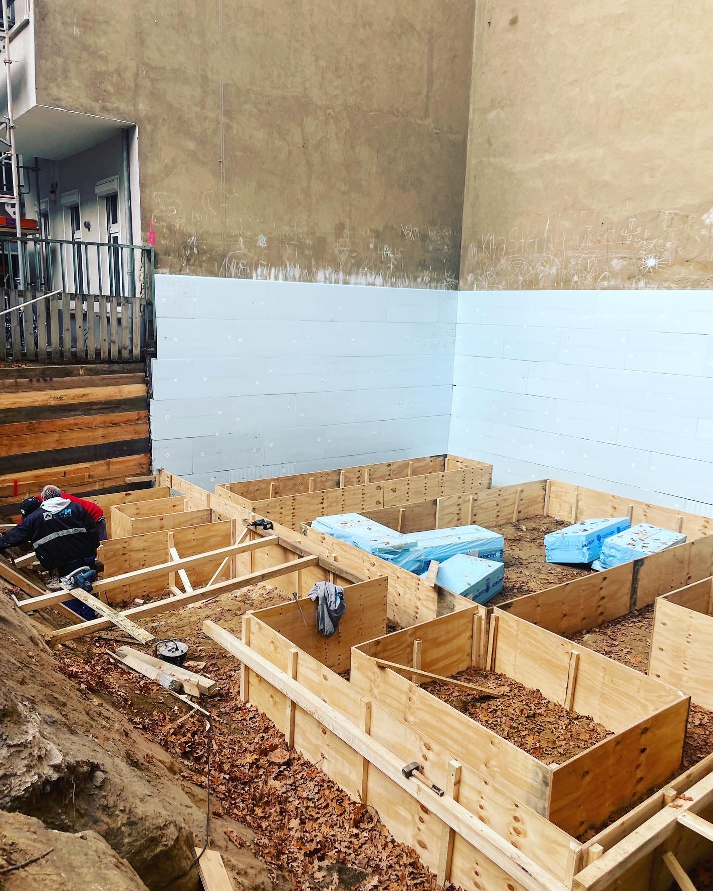 Budowa fundamentów: drewniane szalunki, ocieplenie styropianem, robotnicy w trakcie pracy. Widoczne narzędzia i materiały budowlane.