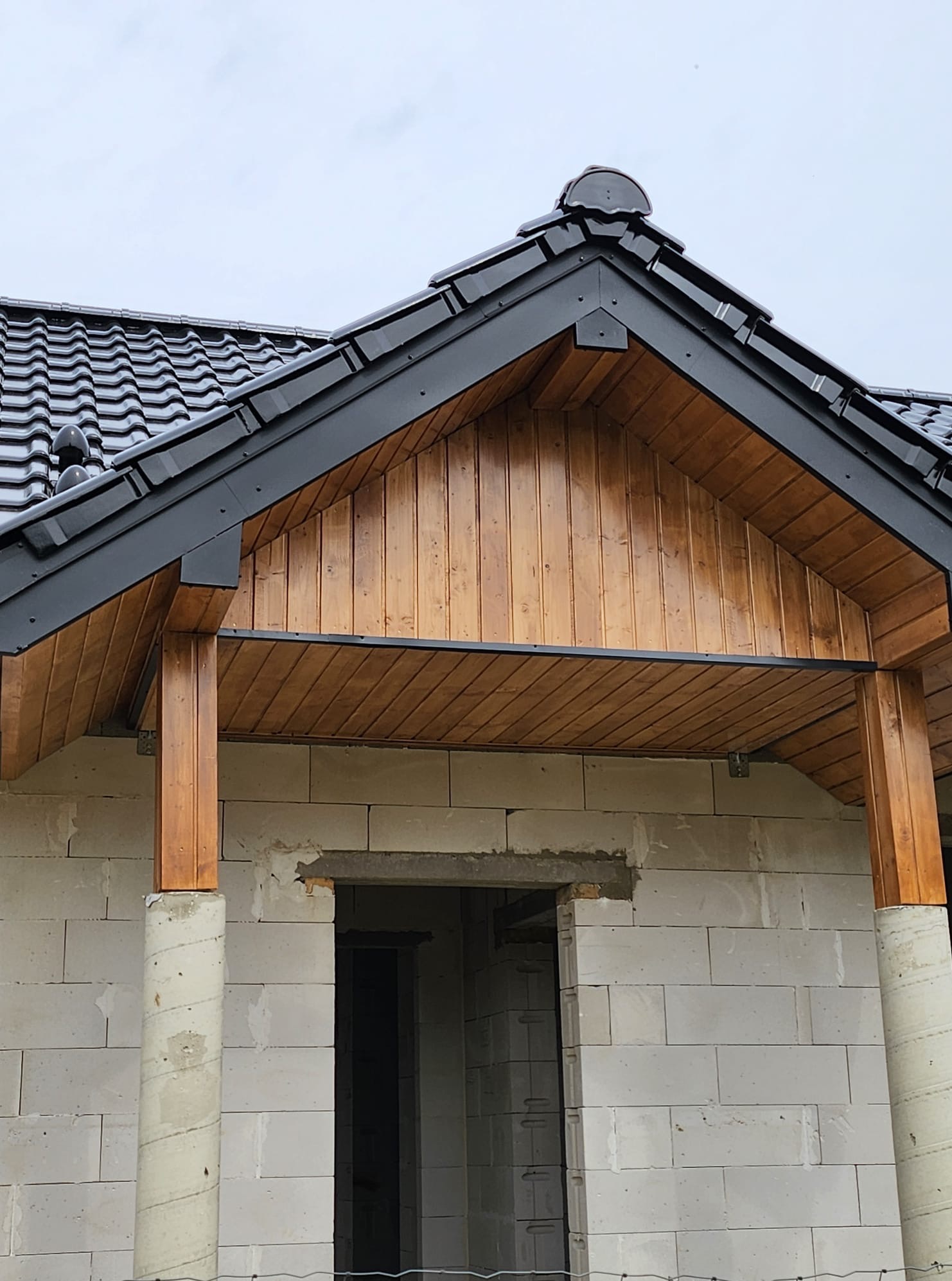 Zadaszenie wejścia do domu w stanie surowym: czarna dachówka, drewniana boazeria i kolumny na ścianie z białego betonu komórkowego.