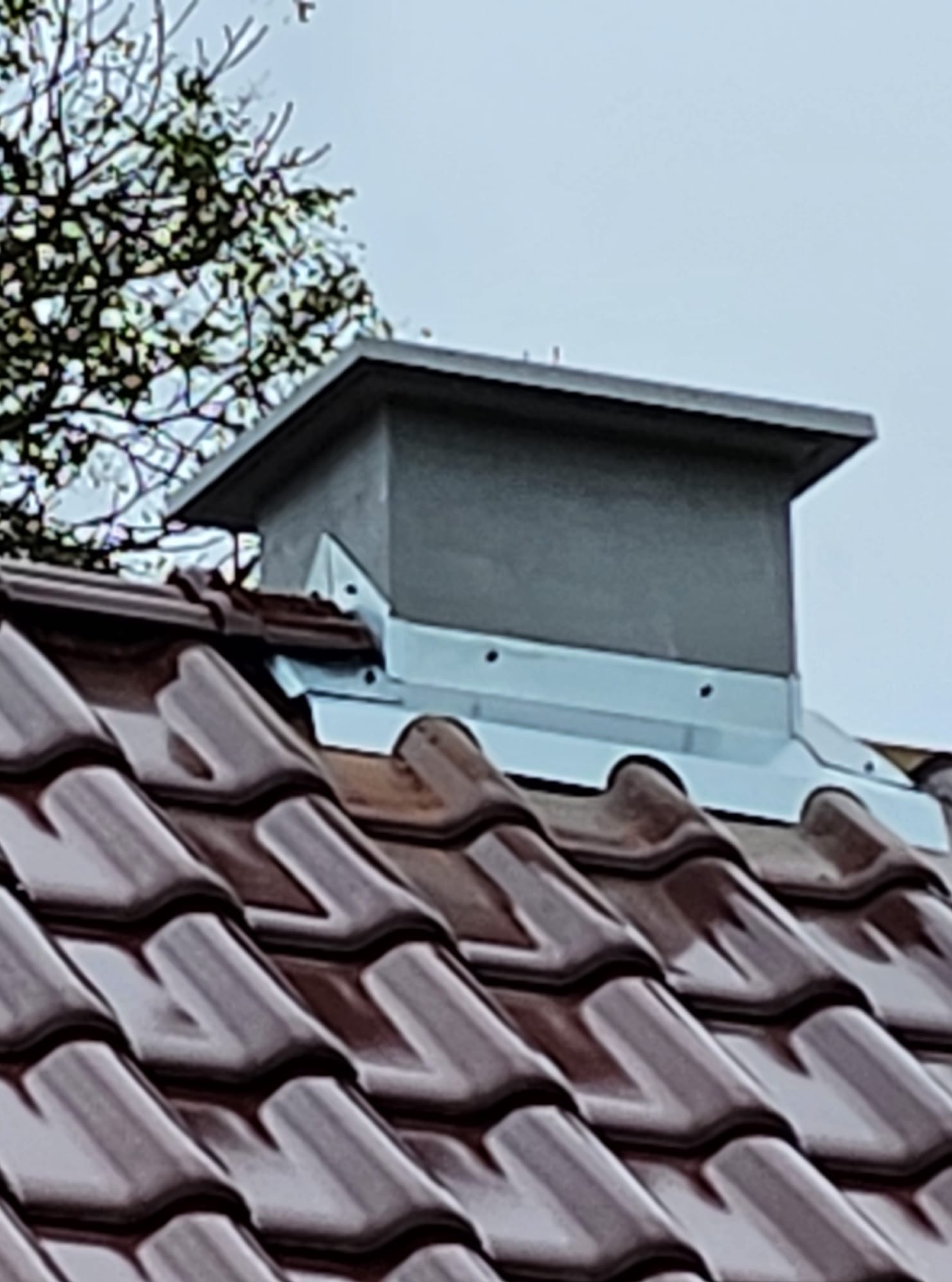 Szczegółowe ujęcie brązowej dachówki ceramicznej z szarym kominem wentylacyjnym, widoczne mocowania blaszane i fragment drzewa w tle.