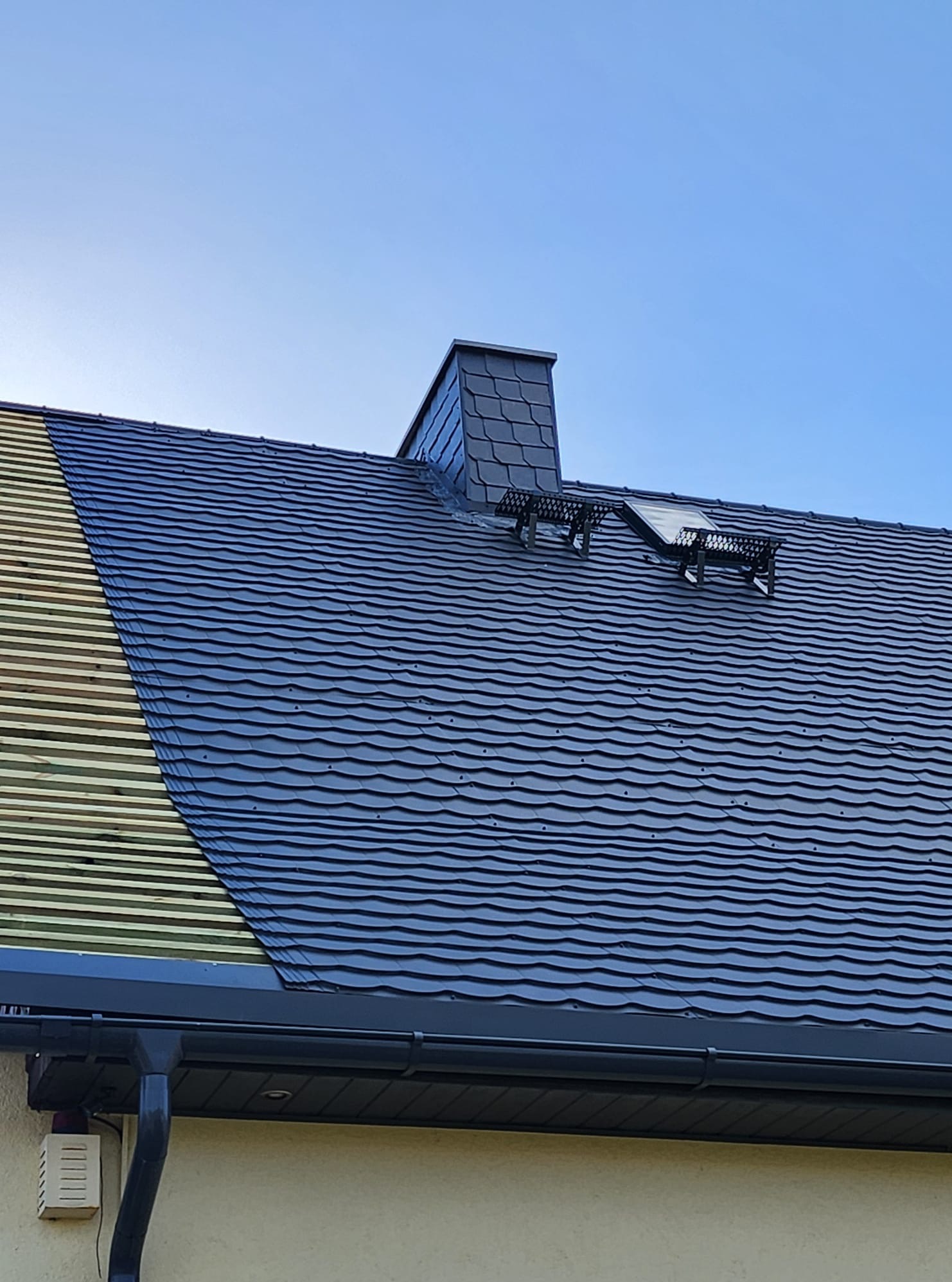 Częściowy widok dachu z czarną dachówką karpiówką i lukarną, fragment drewnianej konstrukcji, orynnowanie. Błękitne niebo w tle.