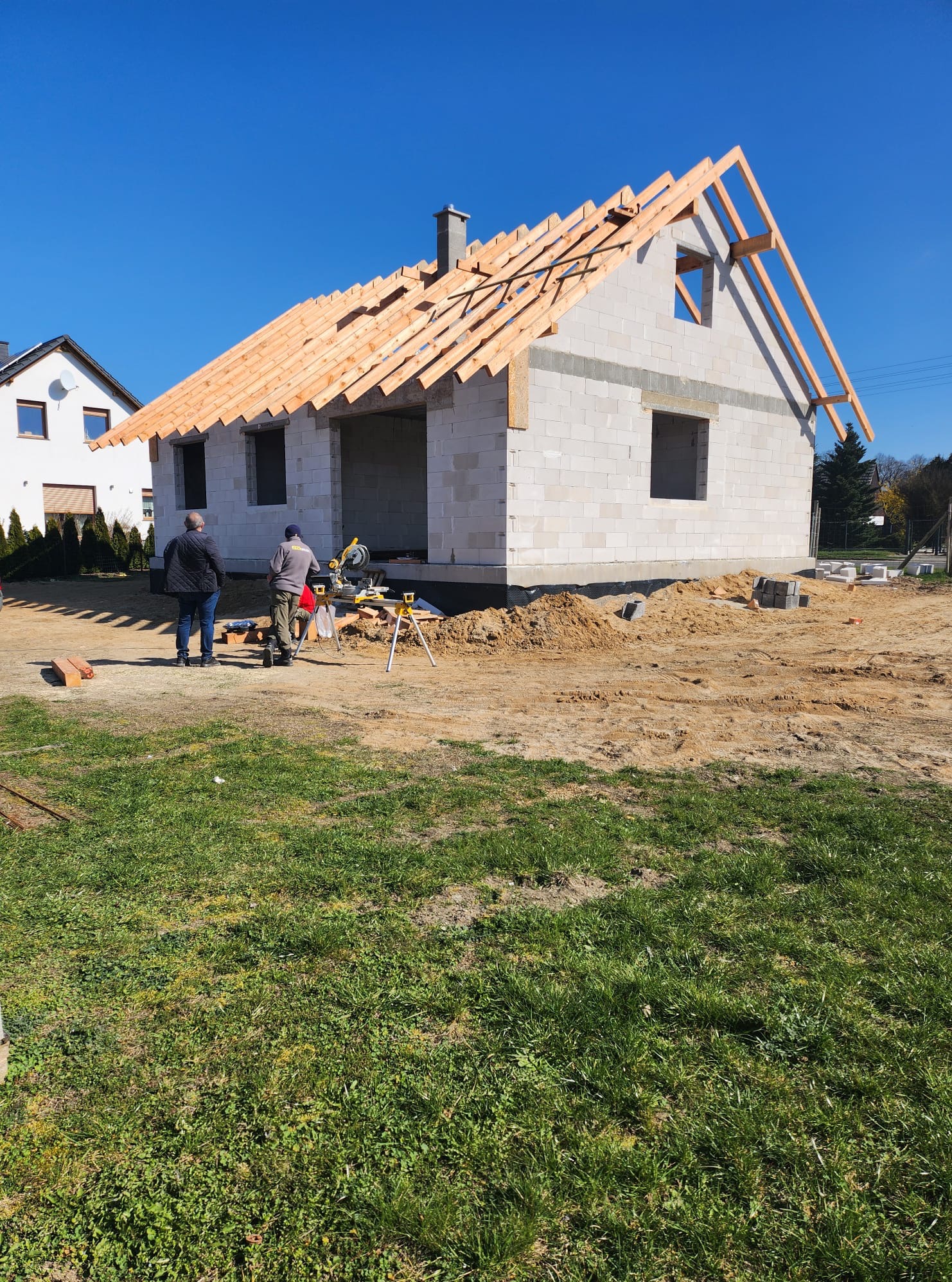 Budowa domu z białych bloczków: ściany wzniesione, więźba dachowa w trakcie montażu. Dwóch pracowników przy pile, przygotowuje elementy konstrukcji dachu.