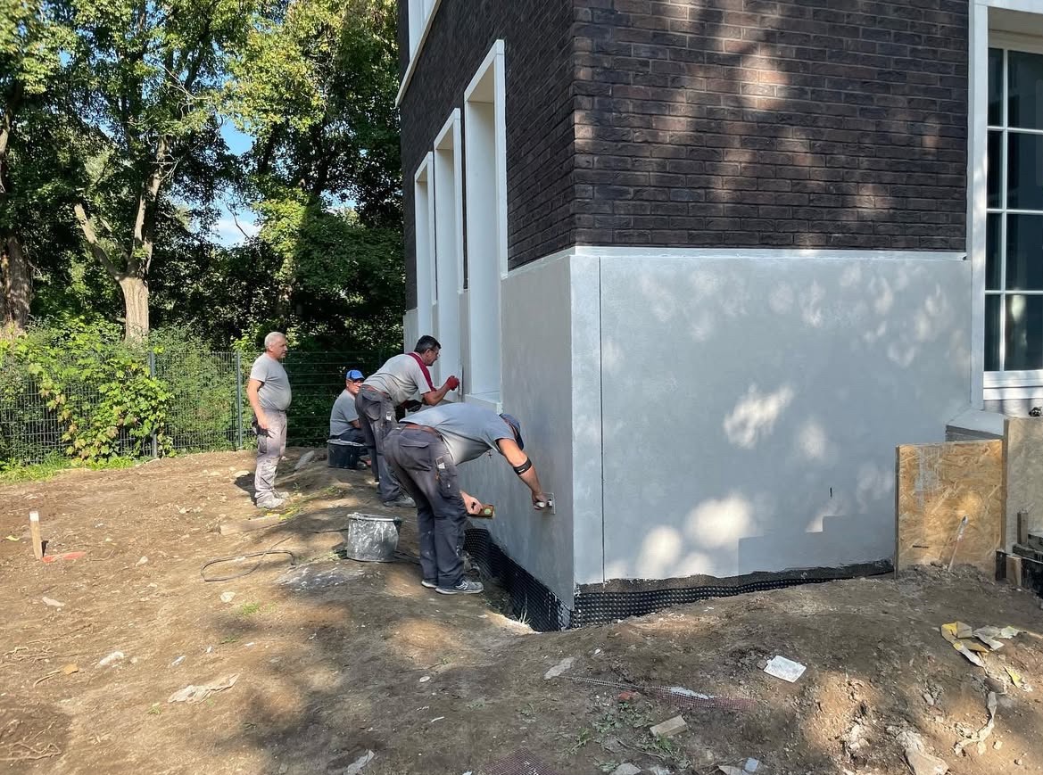 Ekipa tynkarzy wykańcza dolną część elewacji budynku z ciemnej cegły. Widoczne narzędzia i fragment ogrodu. Praca wre, zbliżenie na proces nakładania tynku.