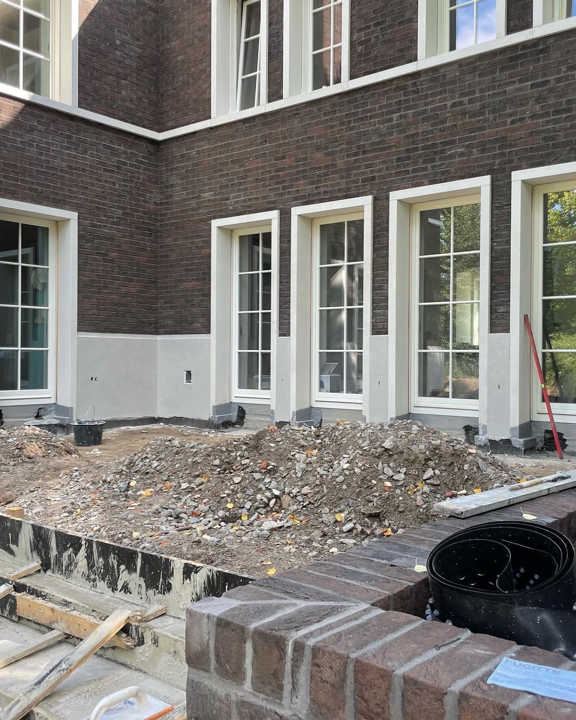 Brunatna ceglana elewacja z białymi oknami w trakcie prac ziemnych. Widoczne narzędzia i gruz. Przygotowanie pod fundament lub taras przy budynku.